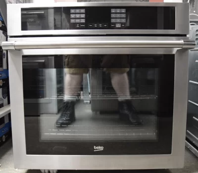 A close-up of a brand new Beko WOS30100SS 30-inch Built-In Single Electric Wall Oven with a reflection of a person's leg on its glass cover.