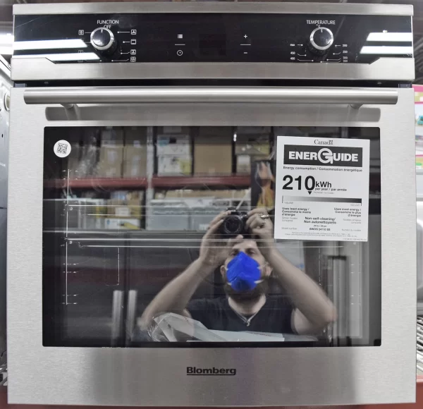 A close-up of a brand new Blomberg BWOS24110SS 24-inch Built-In Wall Oven in stainless steel with a reflection of a photographer holding a camera on its glass.