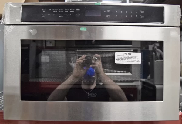 A close-up of a Beko MWDR24100SS 24-inch built-in Microwave Drawer with a reflection of a man holding a camera on its glass.