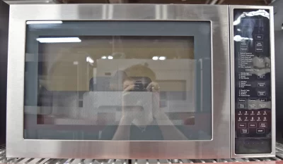 Front view of a brand new Jenn-Air Stainless Steel Series JMC3415ES 25-inch Countertop Convection Microwave Oven with a photographer's reflection on its glass door.
